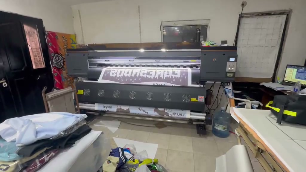 Impresora de gran formato imprimiendo una bandera de egresados, con el diseño en la computadora y prendas apiladas alrededor
